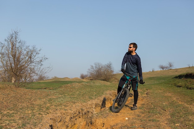 Ciclista em shorts e camisa em uma bicicleta hardtail de carbono moderna com um garfo de suspensão a ar em pé em um penhasco no contexto da floresta verde fresca da primavera