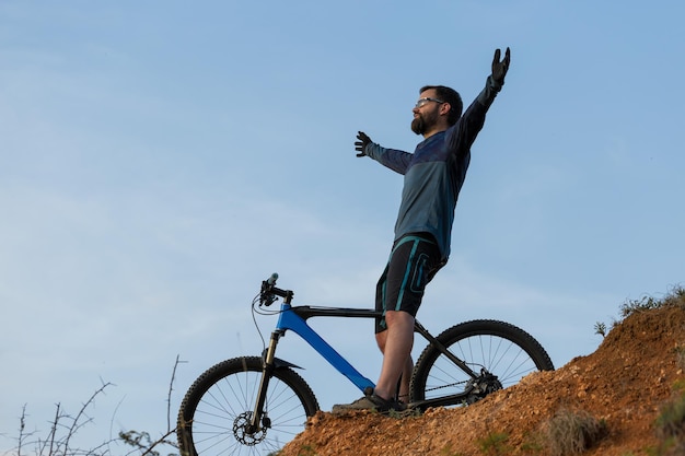 Ciclista em shorts e camisa em uma moderna bicicleta hardtail de carbono com um garfo de suspensão a ar em pé em um penhasco no contexto da floresta verde fresca de verão