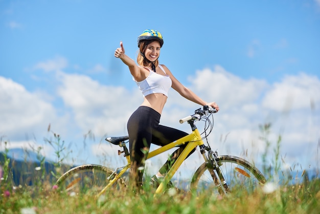 Ciclista femenino sonriente que monta en la bicicleta amarilla en las montañas el día de verano, que muestra los pulgares encima de la muestra contra el cielo azul y las nubes. Actividad al aire libre, concepto de estilo de vida. Copia espacio