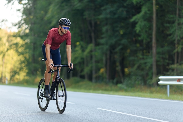 Ciclista guapo feliz en traje deportivo y casco protector montando bicicleta con hermoso bosque verde alrededor