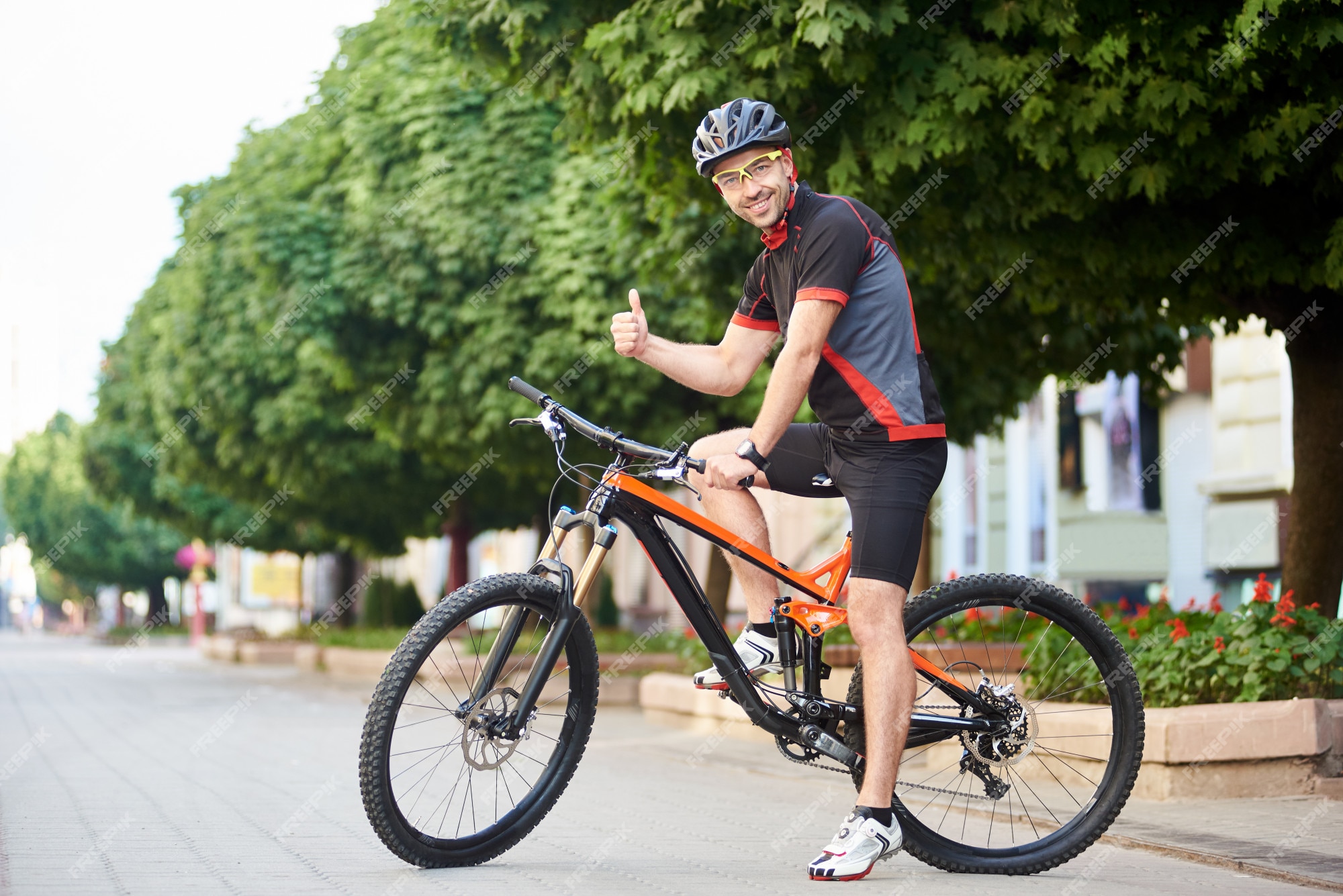 Ciclista de hombre feliz en ropa de ciclismo profesional y ropa protectora mostrando los pulgares sentado en bicicleta a árboles verdes y sonriendo a cámara. deportista satisfecho después del entrenamiento