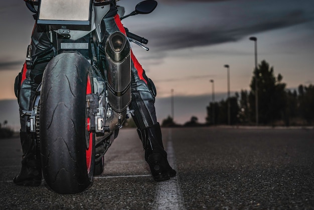 Un ciclista irreconocible sentado en una motocicleta deportiva negra sobre la carretera asfaltada al anochecer bajo un cielo nublado. Copie el espacio.