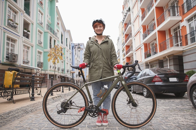 Ciclista jovem e atraente sorrindo para a câmera em pé perto de sua bicicleta na estrada