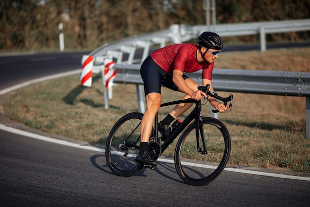 Ciclista joven activo en ropa deportiva y casco protector de carreras en carretera pavimentada durante el día soleado