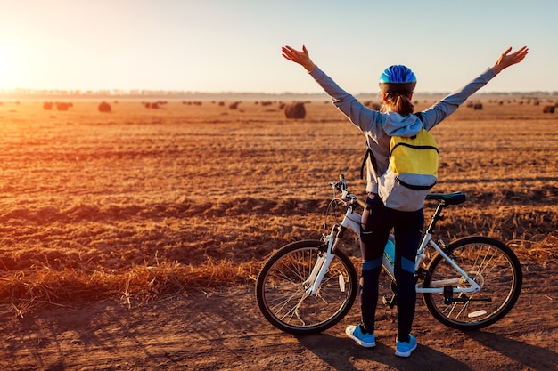 Ciclista joven feliz levantando los brazos abiertos en el campo de otoño admirando la vista. Mujer sintiéndose libre. Celebrando la victoria