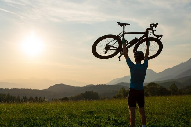 Foto ciclista levantando una bicicleta por encima de su cabeza y mirando la puesta de sol