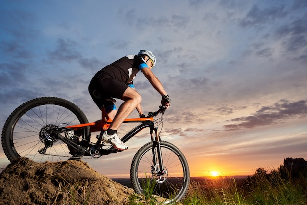 Ciclista masculino activo vistiendo ropa deportiva y casco, montando bicicleta solo y rodando cuesta abajo. Hombre deportivo y robusto en bicicleta contra la hermosa puesta de sol y el cielo azul rosado en la noche