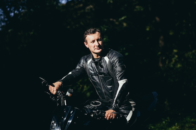 Foto ciclista masculino en una chaqueta de cuero negro sentado en una motocicleta deportiva negra en una carretera forestal