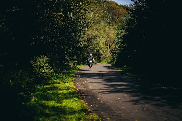 Ciclista masculino en una chaqueta de cuero negro sentado en una motocicleta deportiva negra en una carretera forestal