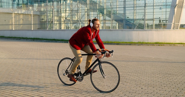Ciclista masculino elegante joven caucásico en chaqueta casual roja y auriculares que andan en bicicleta en la calle en la ciudad. Hombre guapo con andar en bicicleta y escuchar música. Paisaje de la ciudad. Fuera de.