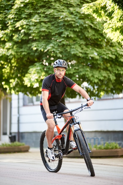 Ciclista masculino joven, mirando a cámara, andar en bicicleta por el callejón vacío del centro de la ciudad rodeado de árboles verdes.