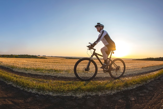 El ciclista monta una bicicleta en la carretera cerca del campo con el telón de fondo del sol poniente. Deportes al aire libre. Estilo de vida saludable.