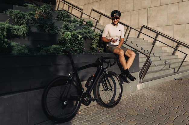 Ciclista muscular profissional sorrindo para a câmera enquanto estiver usando smartphone na rua da cidade bicicleta esportiva preta em pé perto de relaxamento após o treino com gadgets nas mãos