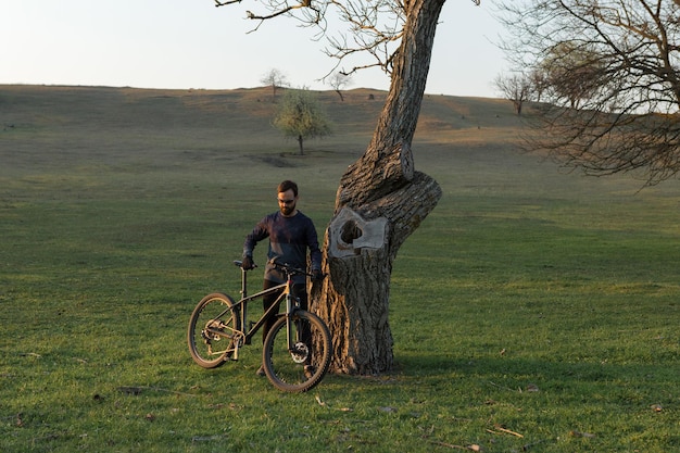 Ciclista en pantalones cortos y jersey en una moderna bicicleta rígida de carbono con una horquilla de suspensión neumática de pie sobre un acantilado con el telón de fondo de un bosque de primavera verde fresco