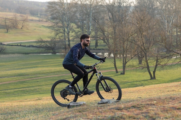 Ciclista en pantalones cortos y jersey en una moderna bicicleta rígida de carbono con una horquilla de suspensión neumática de pie sobre un acantilado con el telón de fondo de un bosque de primavera verde fresco