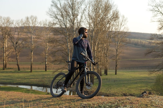 Ciclista en pantalones cortos y jersey en una moderna bicicleta rígida de carbono con una horquilla de suspensión neumática de pie sobre un acantilado con el telón de fondo de un bosque de primavera verde fresco