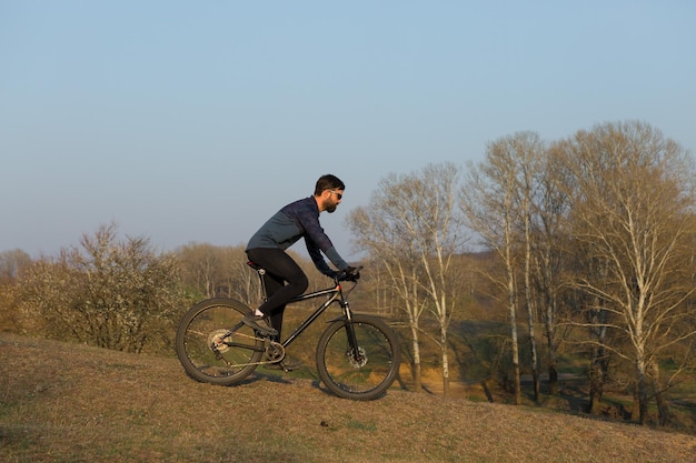 Ciclista en pantalones cortos y jersey en una moderna bicicleta rígida de carbono con una horquilla de suspensión neumática de pie sobre un acantilado con el telón de fondo de un bosque de primavera verde fresco