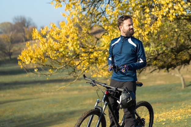 Ciclista en pantalones cortos y jersey en una moderna bicicleta rígida de carbono con una horquilla de suspensión neumática de pie sobre un acantilado con el telón de fondo de un bosque de primavera verde fresco