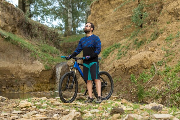 Ciclista en pantalones cortos y jersey en una moderna bicicleta rígida de carbono con una horquilla de suspensión neumática de pie sobre un acantilado con el telón de fondo de un bosque de primavera verde fresco