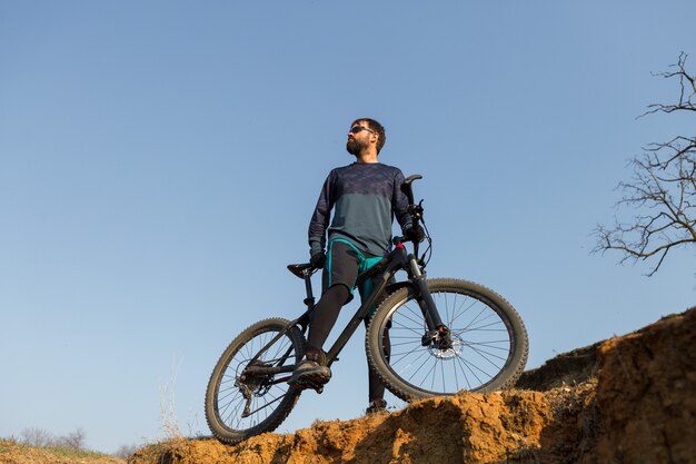 Ciclista en pantalones cortos y maillot en una moderna bicicleta rígida de carbono con una horquilla de suspensión neumática de pie sobre un acantilado con el telón de fondo del bosque de primavera verde fresco