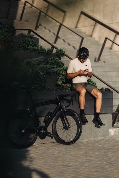 Ciclista con piernas musculosas sentado al aire libre cerca de su bicicleta y usando un teléfono inteligente moderno Hombre caucásico con ropa deportiva, casco y gafas Gadgets modernos y concepto de actividad