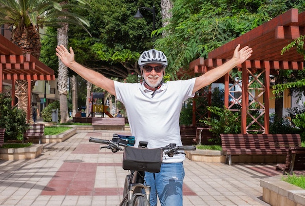 Ciclista senior con casco de pie con los brazos abiertos en el parque público de la ciudad mirando a la cámara sonriendo Concepto de estilo de vida saludable para jubilados y movilidad sostenible