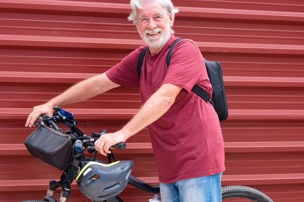 Ciclista senior en excursión con su bicicleta apoyada contra un panel de metal rojo mirando a la cámara