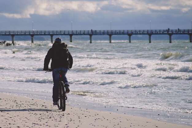 Foto un ciclista solitario con ropa oscura viaja por una playa de arena hacia el muelle