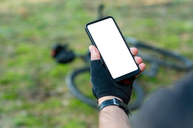 Ciclista sostiene un primer plano de smartphone de maqueta con una pantalla blanca en sus manos en el fondo de una bicicleta en la naturaleza.