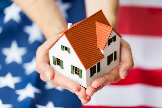 cidadania, residência, propriedade, imóveis e conceito de pessoas - close-up de mãos segurando o modelo de casa viva sobre a bandeira americana