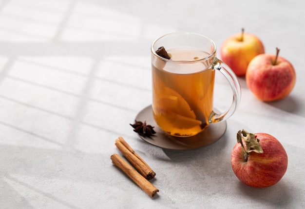 Cidra de maçã caseira com maçãs e canela em copo sobre um fundo claro com frutas frescas, especiarias e sombras O conceito de uma bebida saudável de outono ou inverno