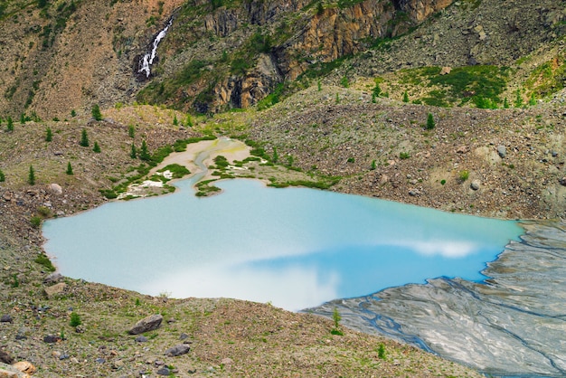 Cielo azul reflejado en el hermoso lago cerca de la ladera de piedra de la montaña. Cascada en la ladera de la montaña. Superficie azul del agua. Arena blanca y grandes piedras en la costa. Paisaje inusual de la naturaleza de Altai.