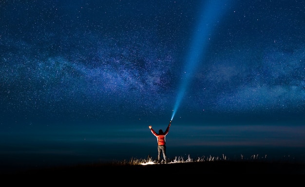 Cielo nocturno con la Vía Láctea y la silueta de un hombre feliz de pie con luz azul clásica.