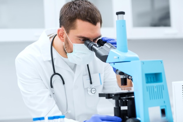 ciência, medicina, tecnologia, biologia e conceito de pessoas - jovem cientista masculino com tubos de ensaio olhando para microscópio e fazendo ou pesquisa em laboratório clínico