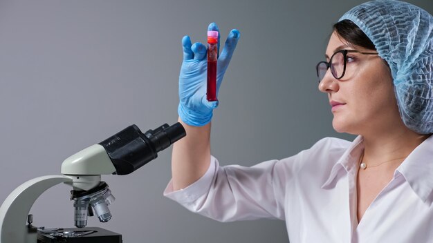 Científica experta con gafas mira una muestra de líquido rojo en un tubo de ensayo cerca del microscopio que trabaja en la vista cercana del hospital moderno