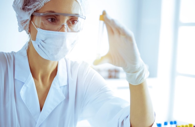 Científica profesional en anteojos protectores investigando tubo con reactivos en un laboratorio soleado tonificado en azul Investigación de medicina y ciencia