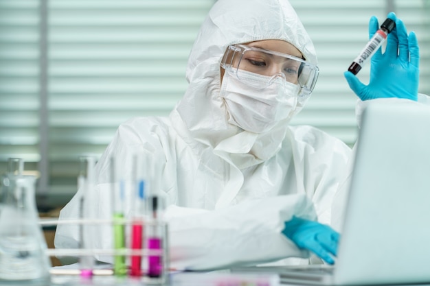 Científica en traje de materiales peligrosos con máscara que sostiene un tubo de muestras de análisis de sangre en la mano escribiendo información del informe sobre el análisis de sangre.