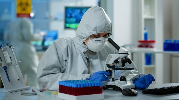 Científica en traje de ppe trabajando en laboratorio con microscopio moderno con portaobjetos. Equipo de investigadores químicos que examinan la evolución del virus utilizando alta tecnología para la investigación científica de vacunas contra covid19