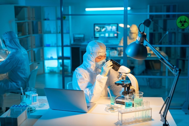 Científica en traje de riesgo biológico respirador y guantes usando microscopio