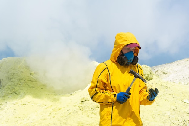 Una científica volcánica en el fondo de una fumarola humeante examina una muestra de un mineral de azufre con un martillo geológico
