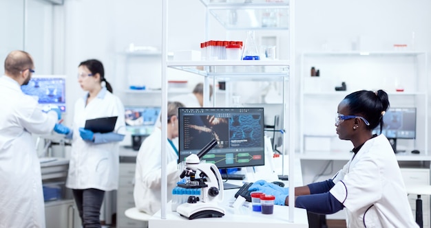 Científico africano sentado en su lugar de trabajo en instalaciones modernas para la industria de la medicina. Investigador de salud negro en laboratorio de bioquímica con equipo estéril.