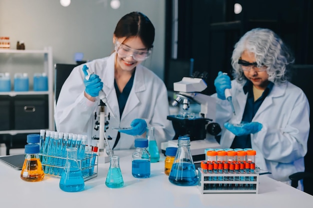 Científico asiático con bata de laboratorio y guantes de protección trabajando con tubos de ensayo con líquidos verdes y rojos con microscopio y otros tubos de ensaio en el fondo en el laboratorio