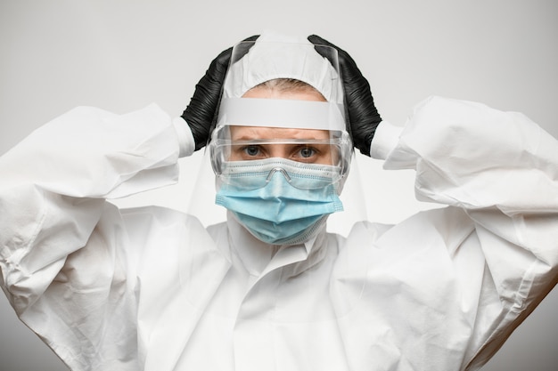 Científico femenino en traje de protección con escudo y máscara médica sostiene sus manos sobre la cabeza.