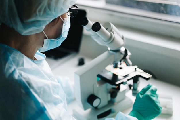 Científico en guantes médicos azules y muestras de aprendizaje uniformes con microscopio en laboratorio