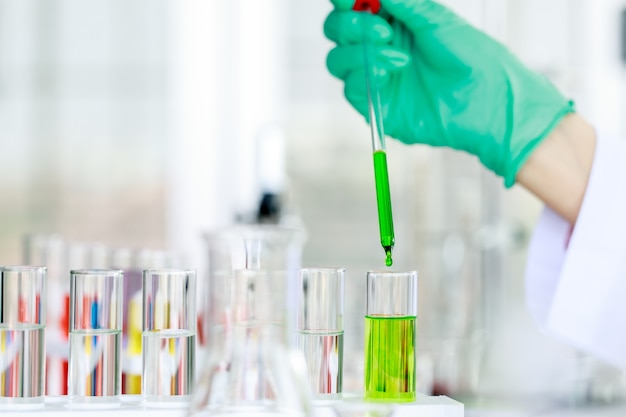 Científico irreconocible de cultivo en bata de laboratorio y guantes con pipeta vertiendo líquido verde en un matraz mientras realiza un experimento químico en el laboratorio.