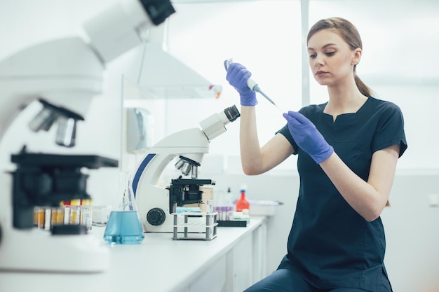 Científico de laboratorio serio sentado en la mesa con un tubo de ensayo en las manos y con cuidado usando un dispensador de pipetas automático