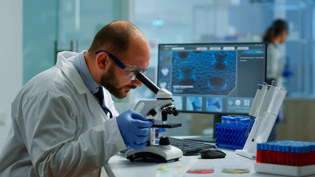 Científico masculino mirando bajo el microscopio en el laboratorio de desarrollo médico, ingresando datos en la computadora. Especialistas que trabajan en medicina, investigación biotecnológica en laboratorio farmacéutico avanzado