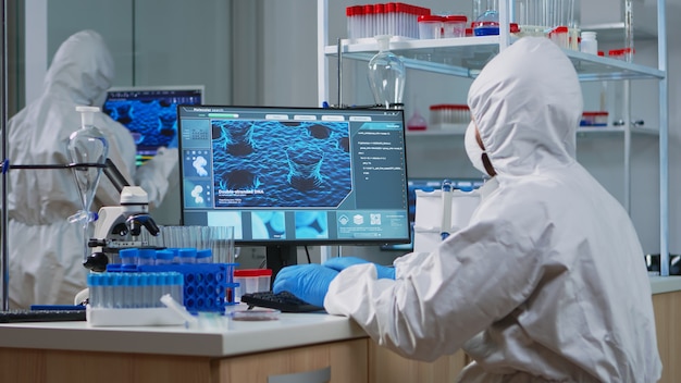 Científico médico en traje de ppe trabajando con imágenes de escaneo de ADN escribiendo en PC en laboratorio equipado. Examinar la evolución de las vacunas utilizando herramientas de alta tecnología y química para el desarrollo de virus de investigación científica