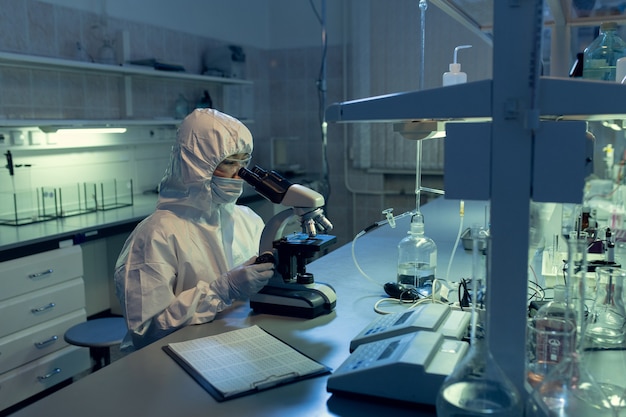 Científico en ropa de trabajo protectora examinando bacterias a través del microscopio en la mesa del laboratorio