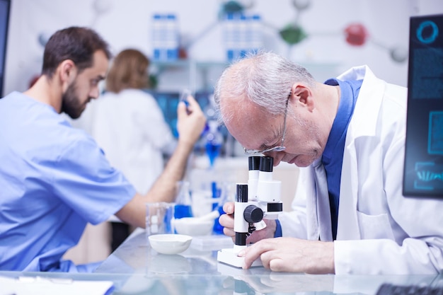 Científico senior con una bata blanca haciendo análisis avanzados bajo el microscopio. Joven científico caucásico en el fondo con un tubo de ensayo.